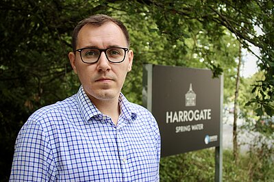 Tom Gordon MP standing next to a Harrogate Spring Water sign, trees in the background.