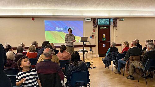 Tom Gordon giving a talk with a powerpoint displaying the Ukraine flag in the background